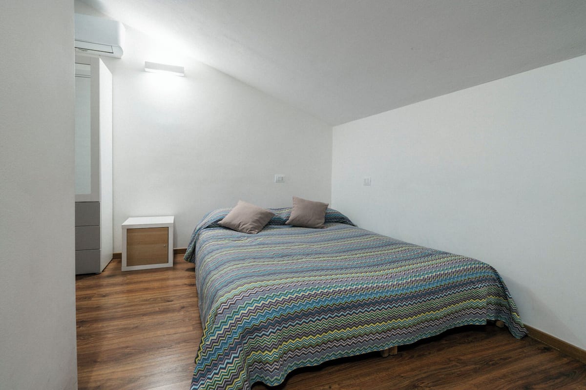 Une petite pièce au mobilier minimaliste, avec un plancher en bois, un lit recouvert d’une courtepointe à motifs zigzag multicolores et une armoire blanche.
