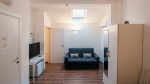 A narrow hallway with a wooden floor, a white door on the left, a small white desk with a television, a dark blue sofa with patterned cushions, and a large wardrobe on the right. - Guest House All'83 (Grosseto)