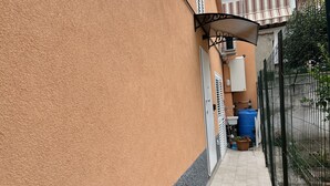 A narrow pathway leading to a building with a textured orange exterior, a metal fence, and a blue barrel. - Guest House All'83 (Grosseto)