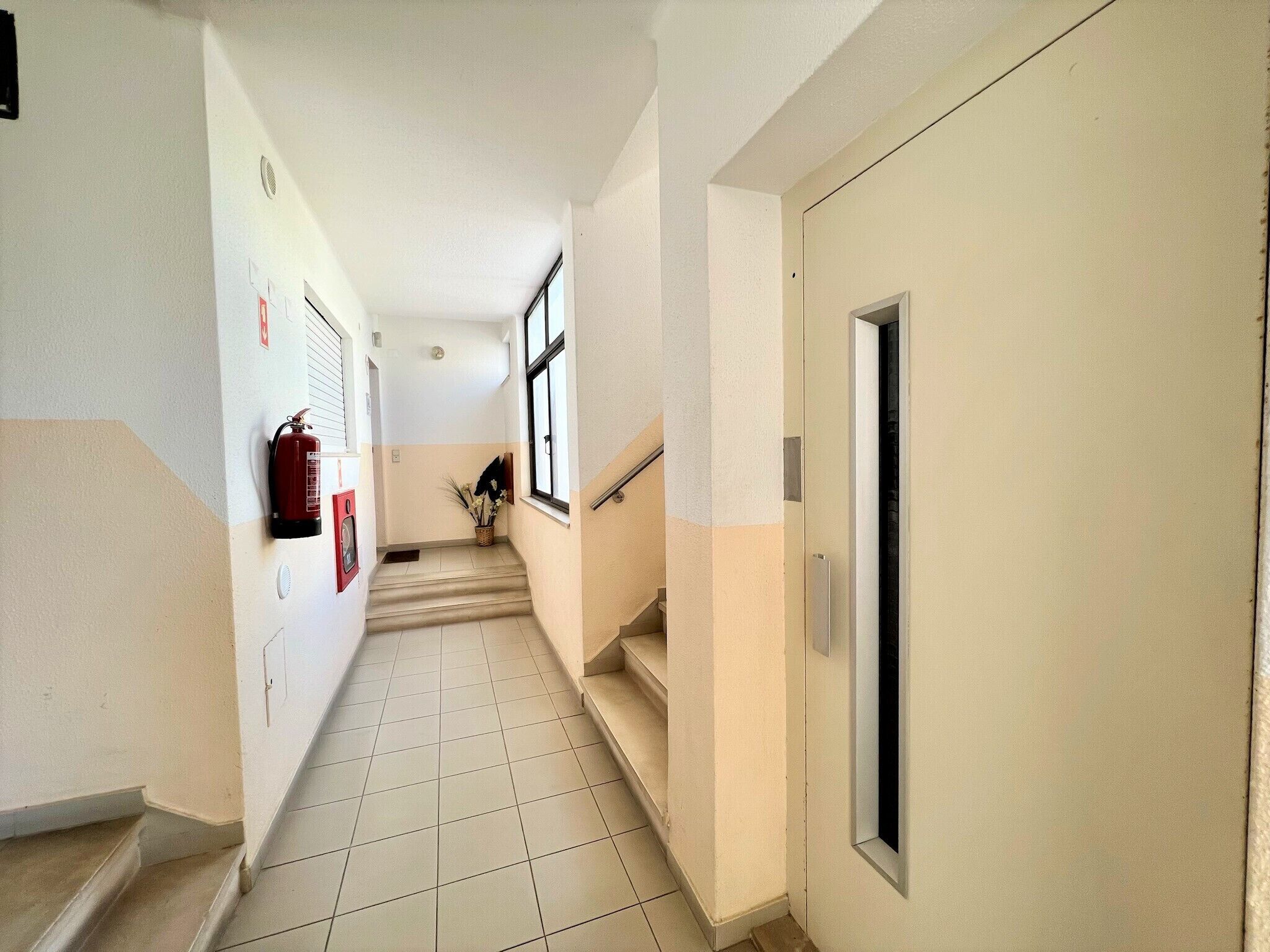 Un couloir doté d’un sol carrelé, d’un extincteur fixé au mur et d’une porte avec une fenêtre.