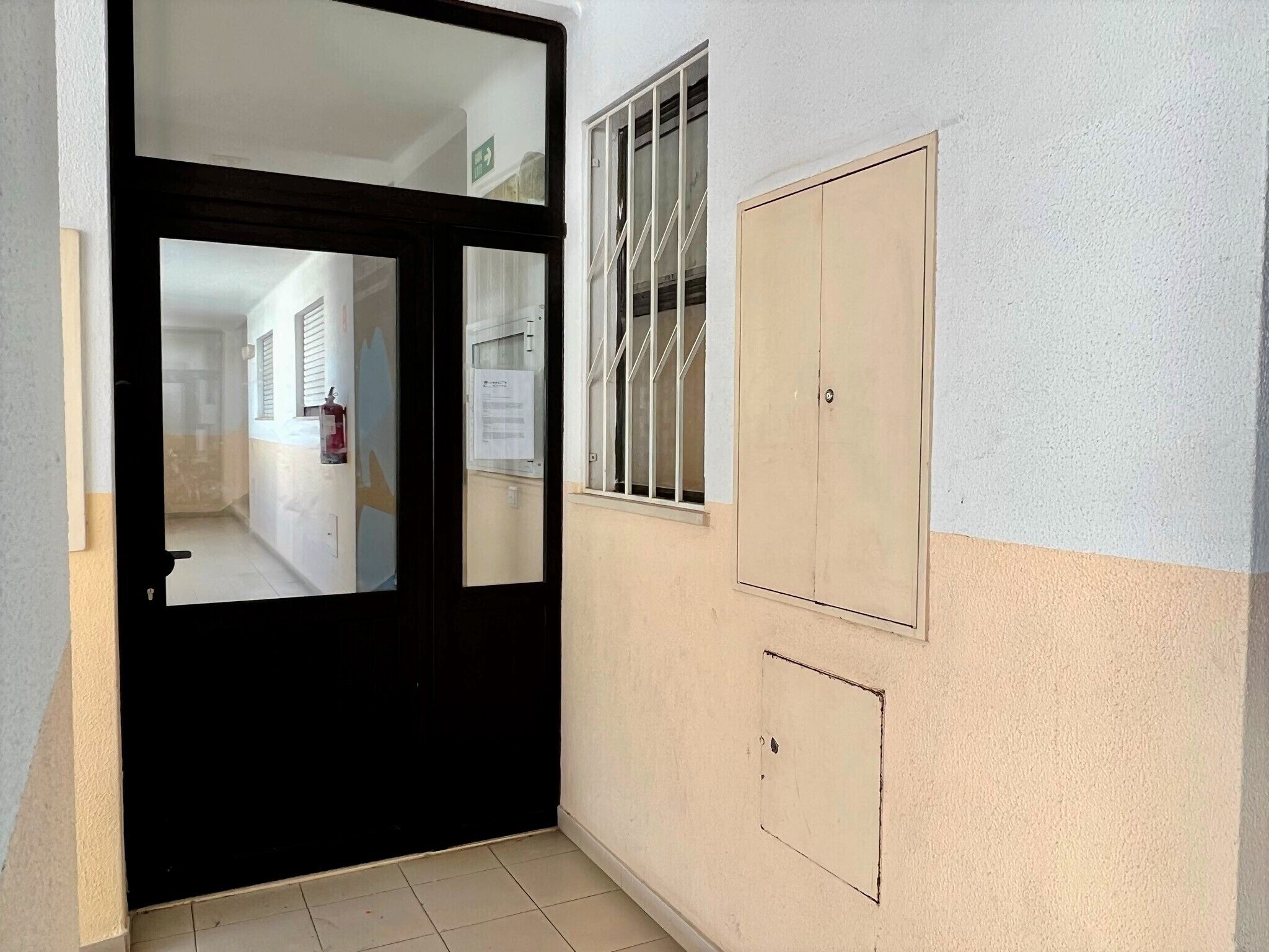 Ein Flur mit einer schwarzen Tür, einem Fenster mit Gitterstäben und einer beigen Wand mit einem kleinen, quadratischen Lüftungsgitter.