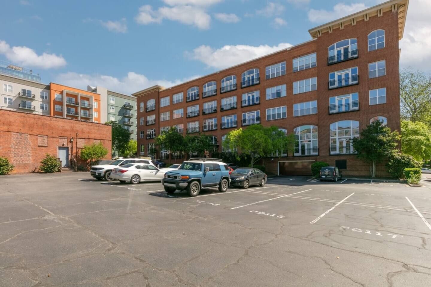 Un parking avec plusieurs voitures, un bâtiment en briques doté de nombreuses fenêtres et de balcons, et un ciel dégagé.
