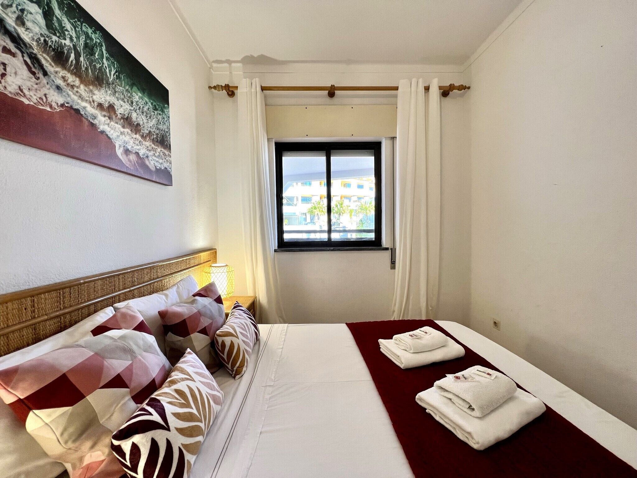 A hotel room with a bed, pillows, a window with curtains, and a painting on the wall.