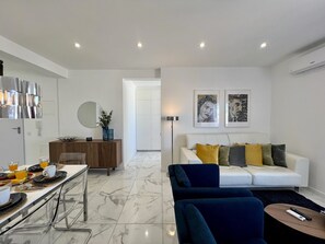 A modern living room with a white sofa, blue armchairs, and a coffee table. There is a dining area with a table set for two, a mirror, and a decorative vase.