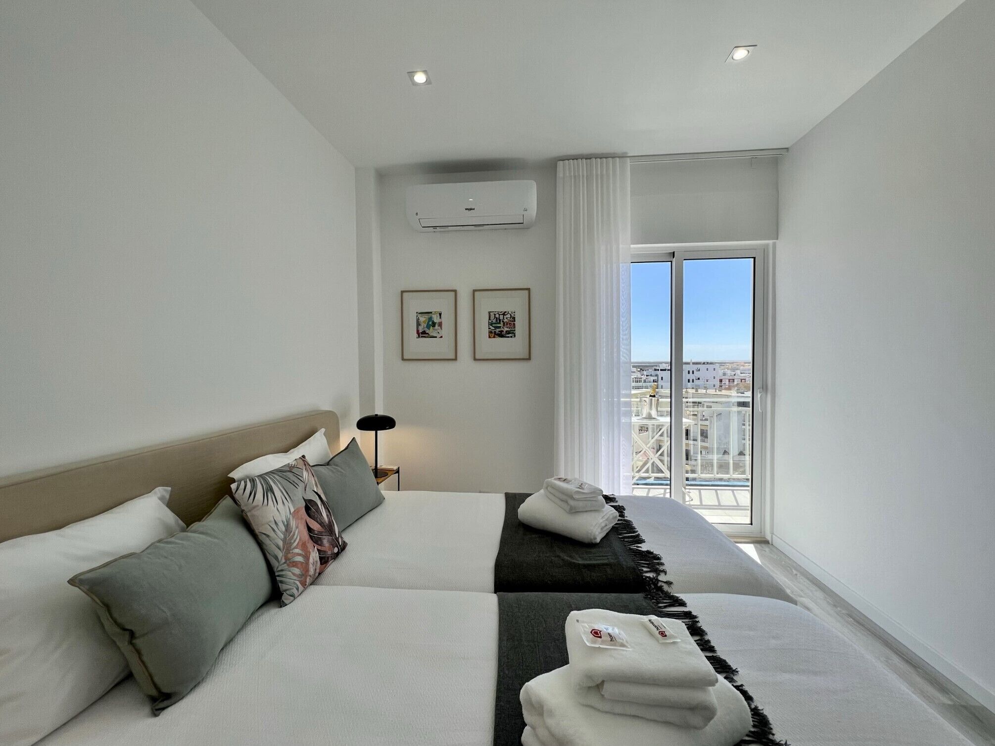 A modern hotel room with a large bed, a view of the city through a large window, and a ceiling-mounted air conditioning unit.