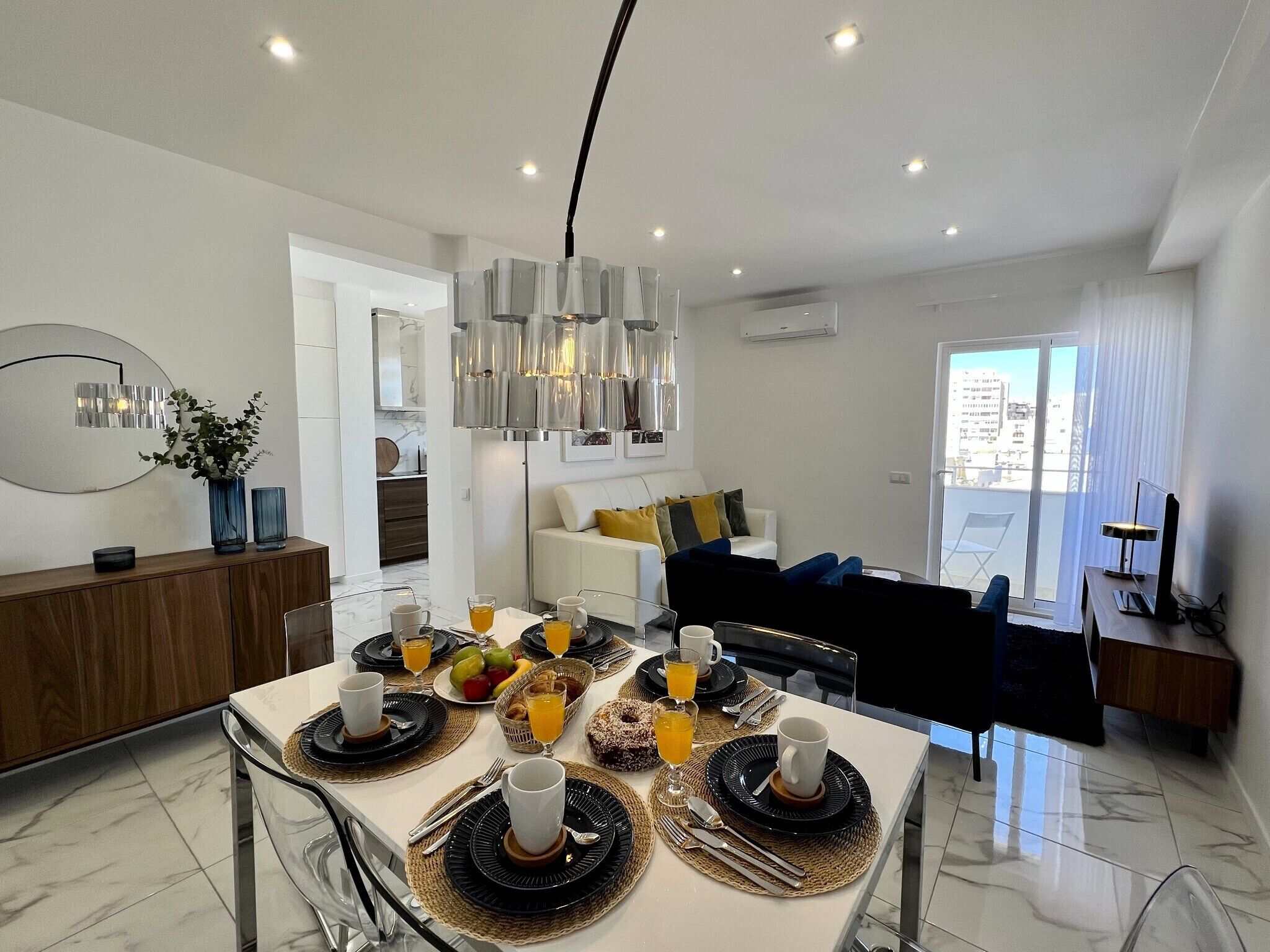 A modern dining area with a white table set for breakfast, a sofa in the background, and a city view through the window.
