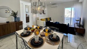 Un comedor moderno con una mesa blanca dispuesta para el desayuno, un sofá al fondo y vistas a la ciudad a través del ventanal.