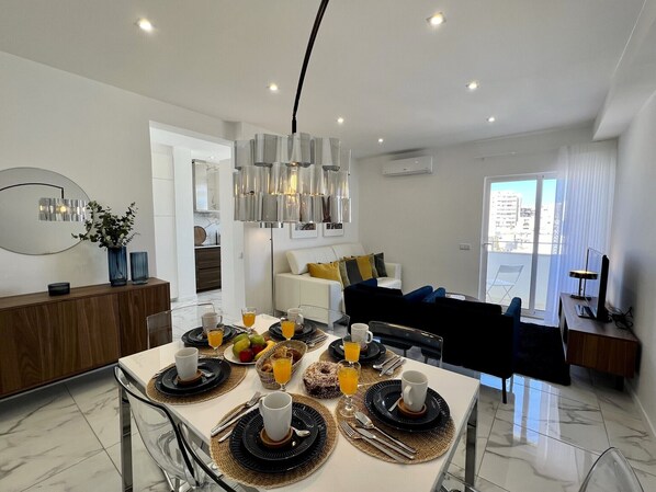 A modern dining area with a white table set for breakfast, a sofa in the background, and a city view through the window.