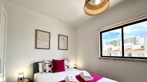 A hotel room with a bed, a nightstand, a lamp, two framed artworks, and a window with a city view. - ALBUFEIRA DESIGN by HOMING (Albufeira)