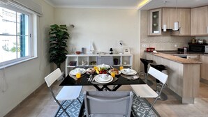 A modern dining area with a black table set for four, surrounded by white chairs. A large potted plant is on the left, and a small white shelf with decorative items is against the wall. - CARVOEIRO TERRACE by HOMING (Carvoeiro)