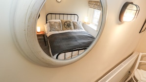 A bedroom with a circular mirror reflecting a bed, a nightstand, and a window with curtains. - Cherry Tree Cottage (Bingley)
