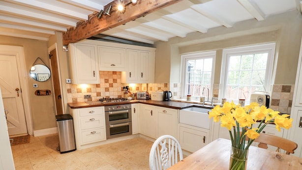 A kitchen with white cabinets, a wooden table with a vase of yellow flowers, and a window with a view of a fence. - Cherry Tree Cottage (Bingley)