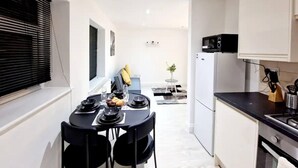 A modern kitchen with white cabinets, a black countertop, and a microwave. There is a dining area with a black table and chairs, and a small plant on a side table. - Modern Apartment in Uxbridge (Uxbridge)