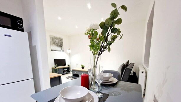 A modern kitchen with a dining table set for two, a vase with flowers, and a flat-screen TV in the background. - Modern Apartment in Uxbridge (Uxbridge)
