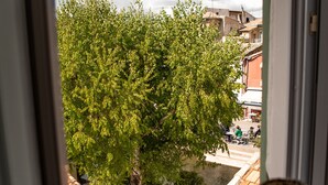 Une vue depuis une fenêtre donnant sur un arbre, un petit espace extérieur aménagé avec des sièges et un bâtiment doté d’un toit en tuiles.