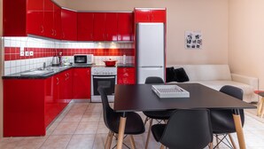 A compact kitchen with red cabinets, a white refrigerator, a microwave, and a stove. There is a dining table with chairs and a small sofa in the living area.