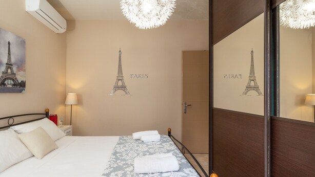 A neatly made bed with a patterned bedspread, a wall-mounted air conditioner, and a large mirror reflecting the room.