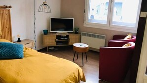 Ett sovrum med en säng, en tv-apparat placerad på ett träskåp, en röd stol och ett fönster med utsikt över byggnader.