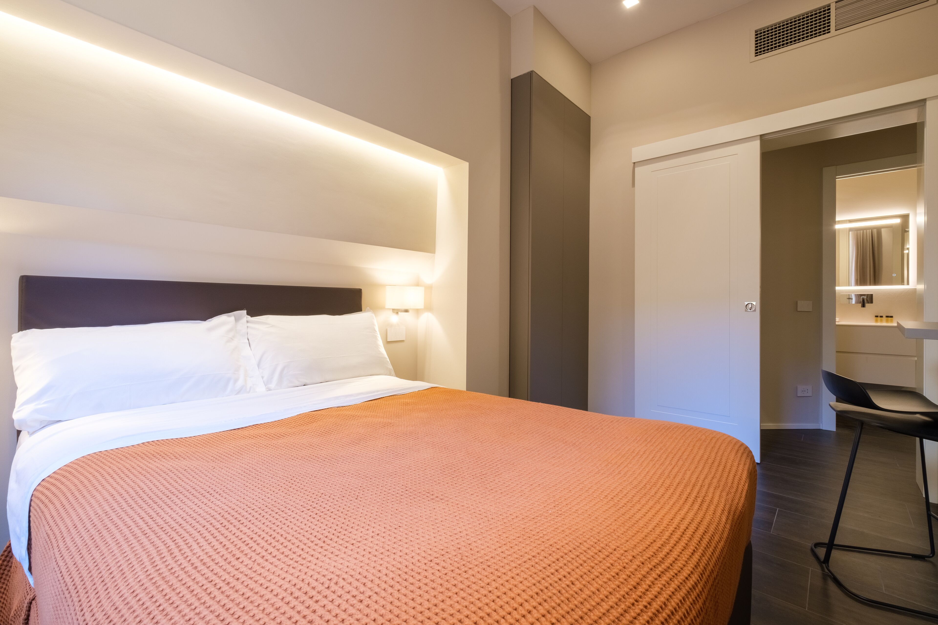 Une chambre d’hôtel moderne avec un grand lit, une table de chevet avec une lampe et une salle de bain visible à travers une porte ouverte.