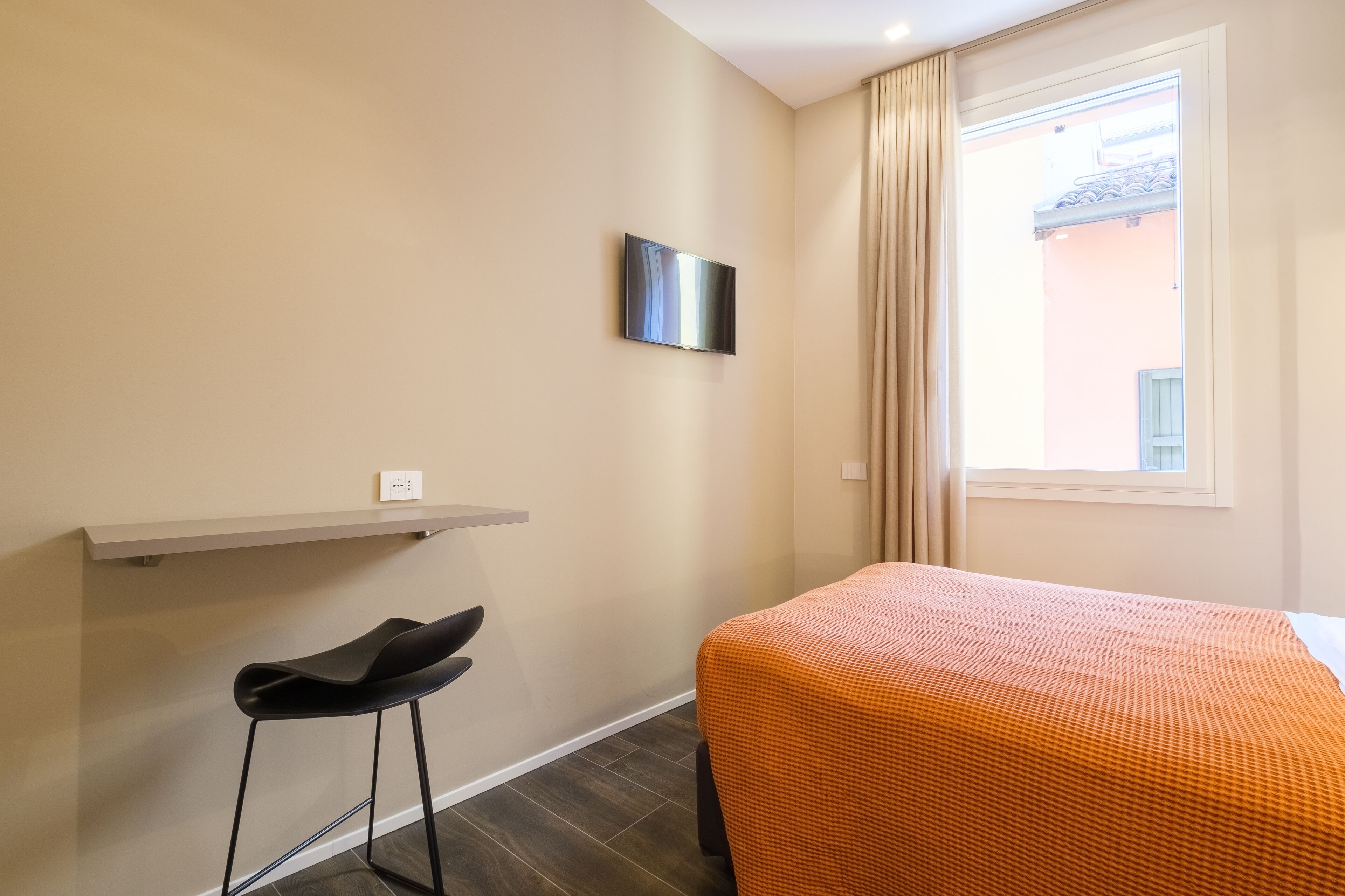 A hotel room with a bed, a TV mounted on the wall, a small stool, and a window with curtains.