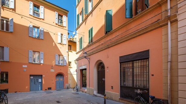 A narrow street with buildings on either side, featuring a mix of closed and open shutters, a bicycle parked on the sidewalk, and a cobblestone street. - Golden Suite by Studio Vita (Bologna)