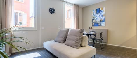 A modern living room with a sofa, a dining area with a table and chairs, and a large window with curtains.