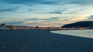 A pebbled beach with a cityscape in the background during twilight. - Luxury Apartment close to the Beach (Llandudno)