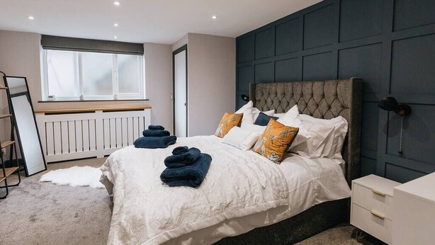 A modern bedroom with a large bed, a dark headboard, and a window with blinds. - Luxury Apartment close to the Beach (Llandudno)