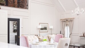 A dining area with a white table set for four, surrounded by upholstered chairs. A chandelier hangs from the ceiling, and a window provides natural light.