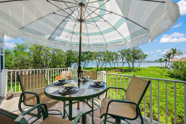 A round table with chairs and an umbrella on a balcony overlooking a body of water and greenery.