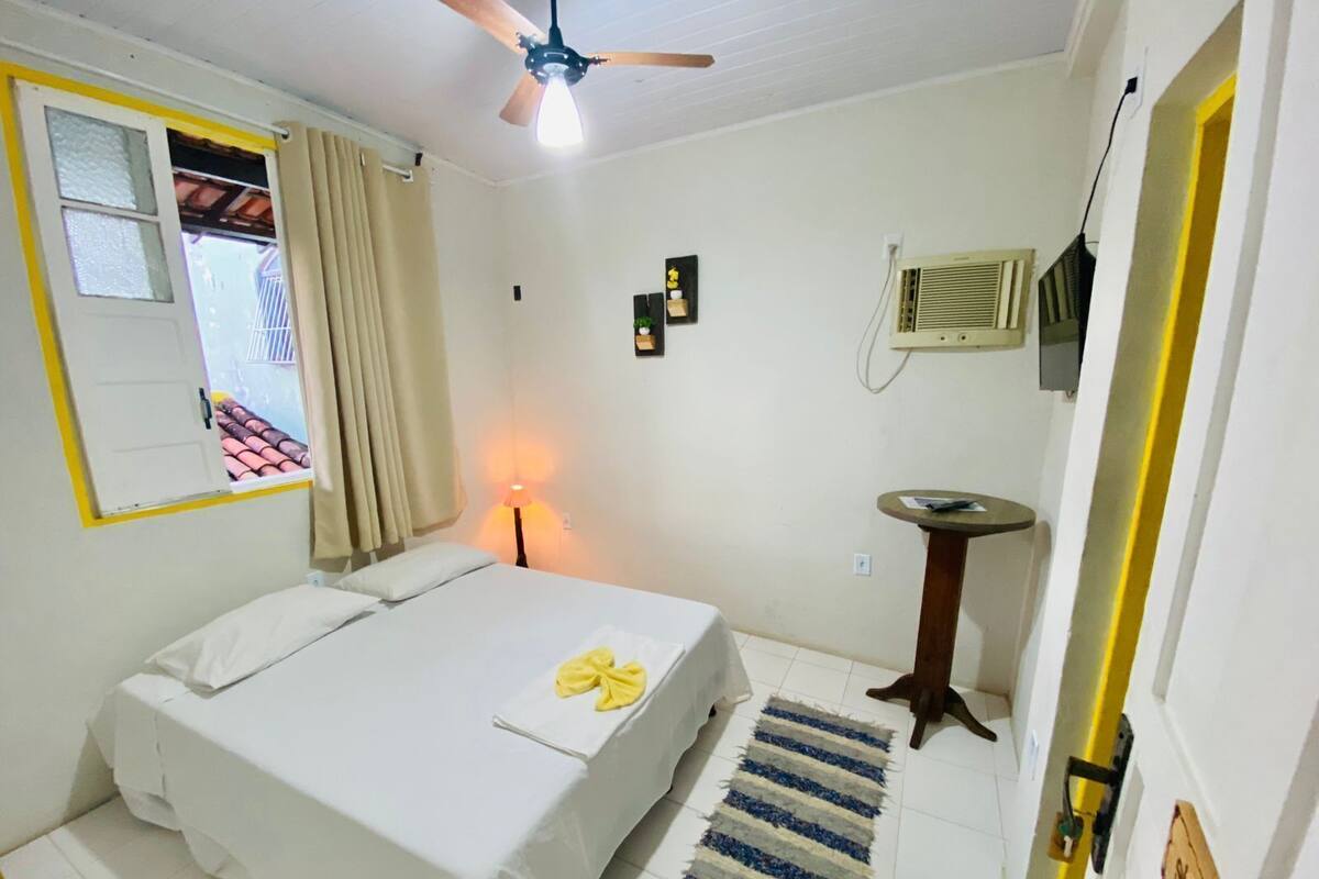 A hotel room with a bed, a ceiling fan, a bedside lamp, a small table, a radiator, and a window with curtains.
