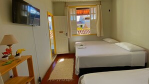 A small, clean room with two beds, a TV, a lamp, and a window with a view of buildings. - Pousada do Beco (Caravelas)
