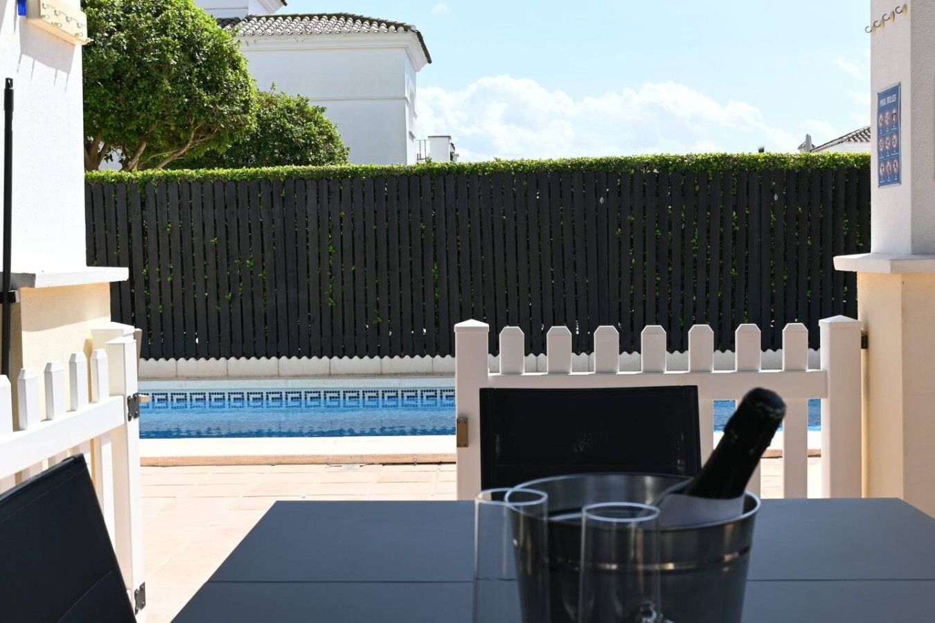 Zona junto a la piscina con valla blanca, piscina y una mesa con vasos.