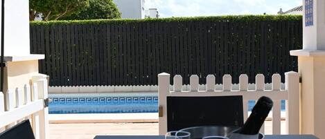 Zona junto a la piscina con valla blanca, piscina y una mesa con vasos.