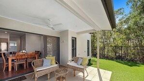 A patio with wicker furniture, a dining area with wooden chairs and a table, and a view of a green lawn and trees. - Private Mudjimba Beach Abode (Mudjimba)