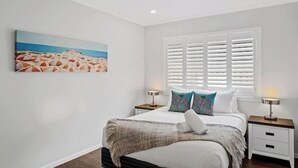 A bedroom with a bed, two bedside tables, a window with shutters, and a beach-themed painting on the wall. - Private Mudjimba Beach Abode (Mudjimba)