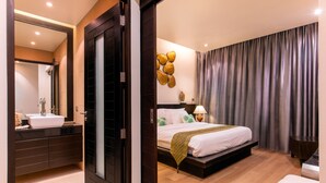Quarto de hotel moderno com piso de madeira, uma cama grande com manta verde, mesa de cabeceira com luminária e banheiro com espelho grande e pia branca.