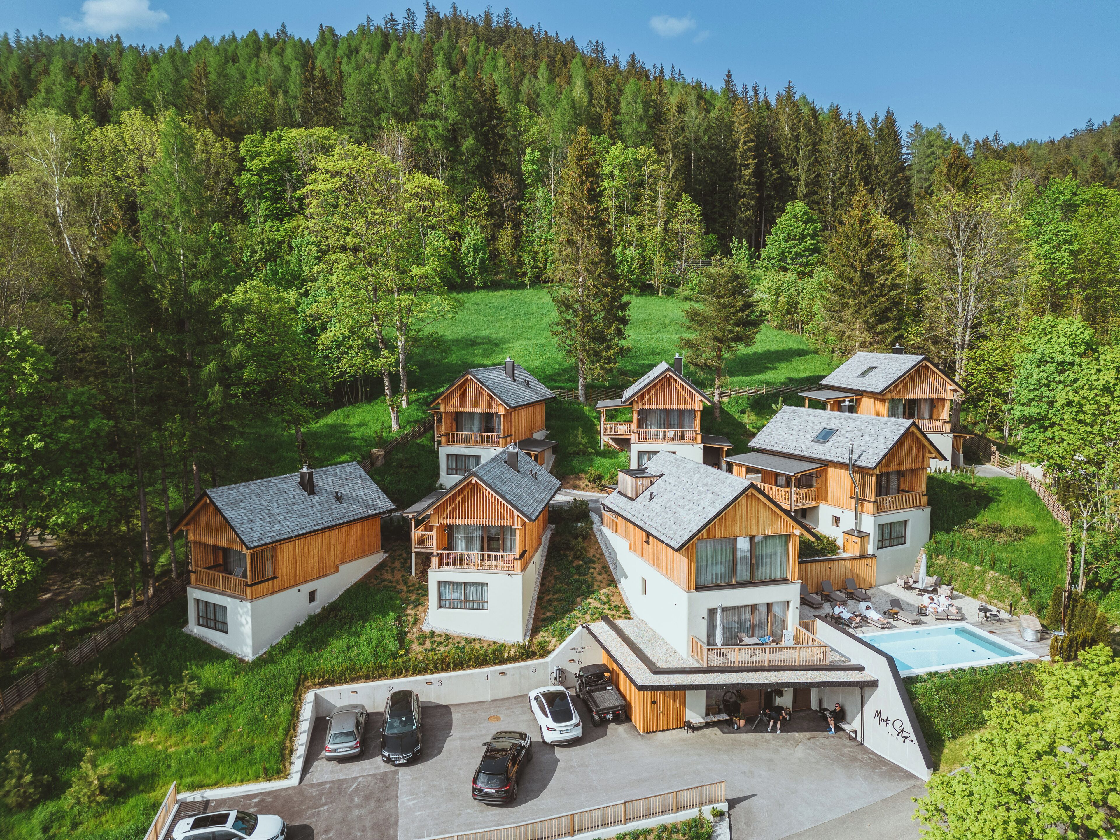 A cluster of wooden cabins with balconies, a swimming pool, and a parking area with cars.