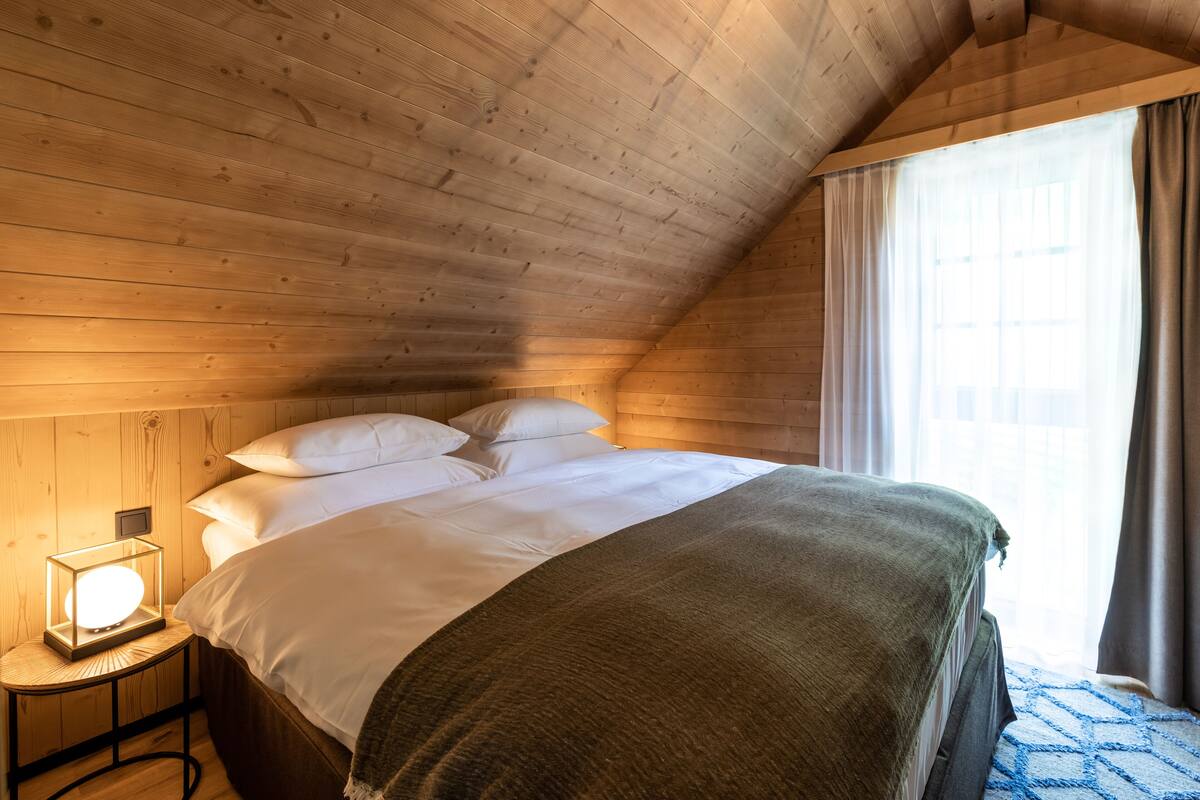 Una habitación con paneles de madera, una cama, una lámpara de noche y una ventana con cortinas.