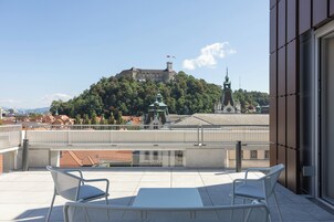 A terrace with white metal chairs and a table, overlooking a hill with a castle and church.
