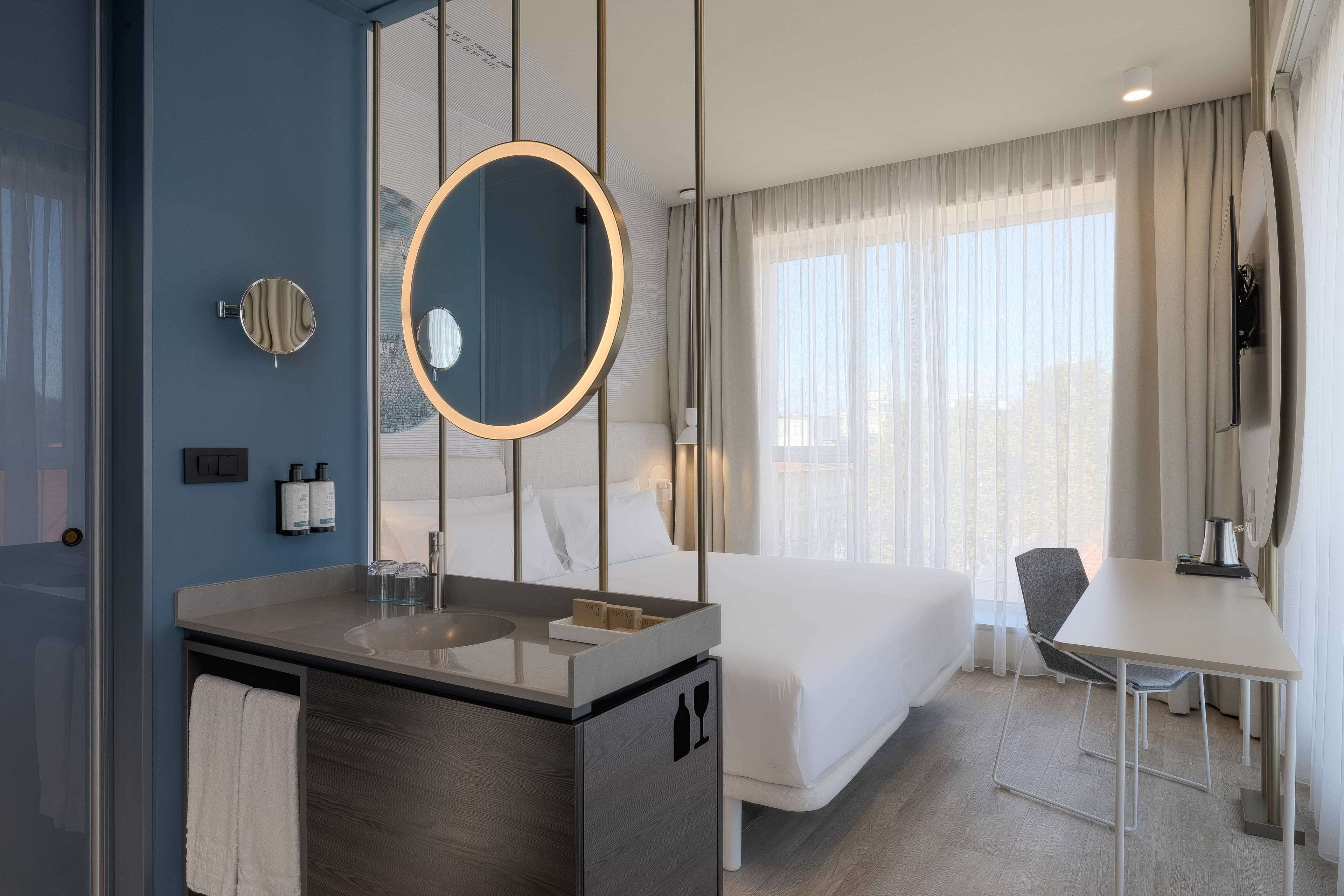 Una habitación de hotel moderna con una cama grande, un escritorio, un espejo redondo y un baño con lavamanos y toalla.