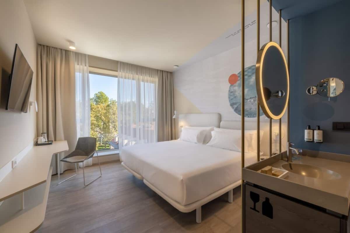 Ein modernes Hotelzimmer mit einem großen Bett, einem Schreibtisch, einem Sessel und einem Badezimmer mit Spiegel, mit Blick ins Freie.