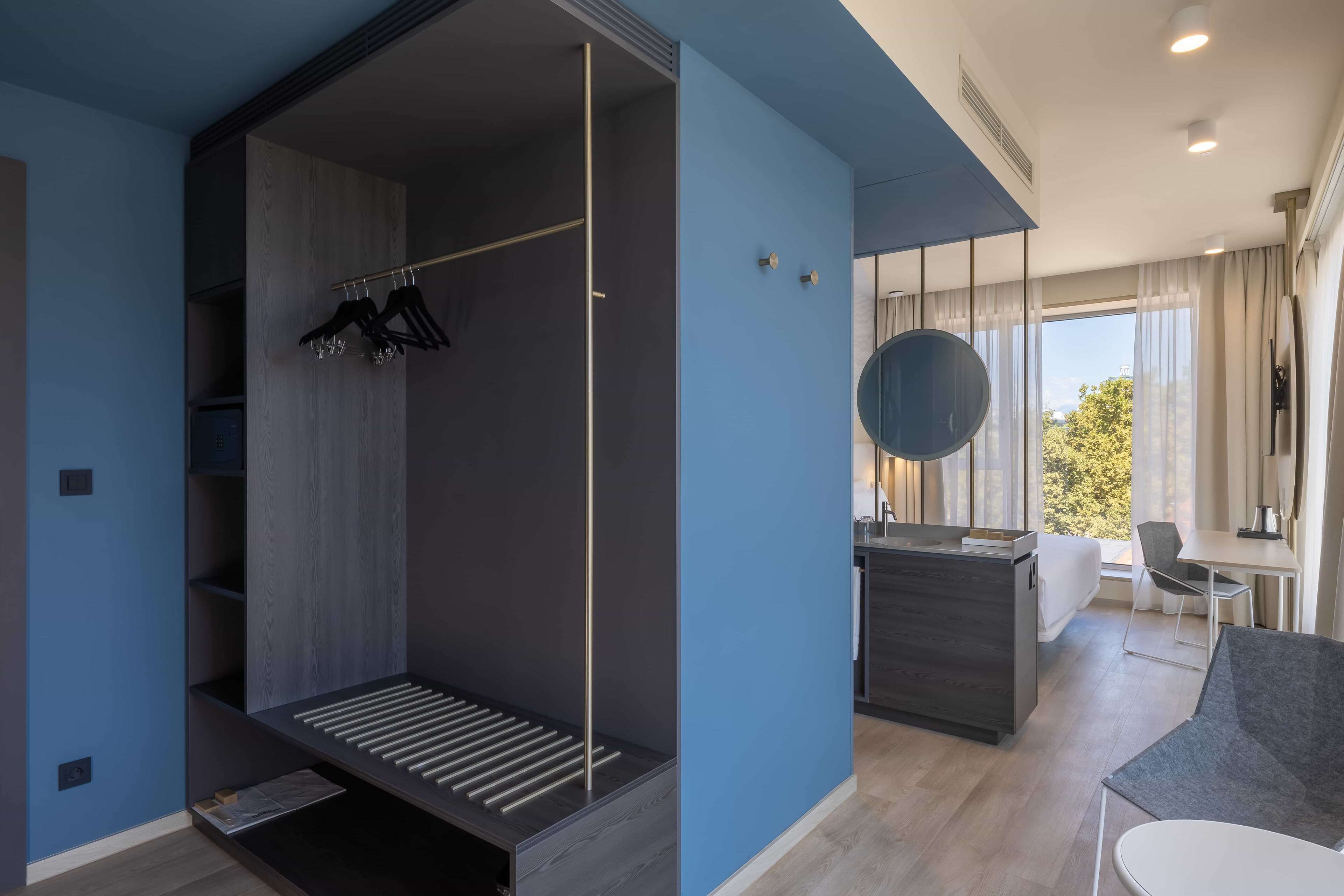 A modern hotel room with a built-in wardrobe, a seating area, a dining table, and a large window offering a view of the outdoors.