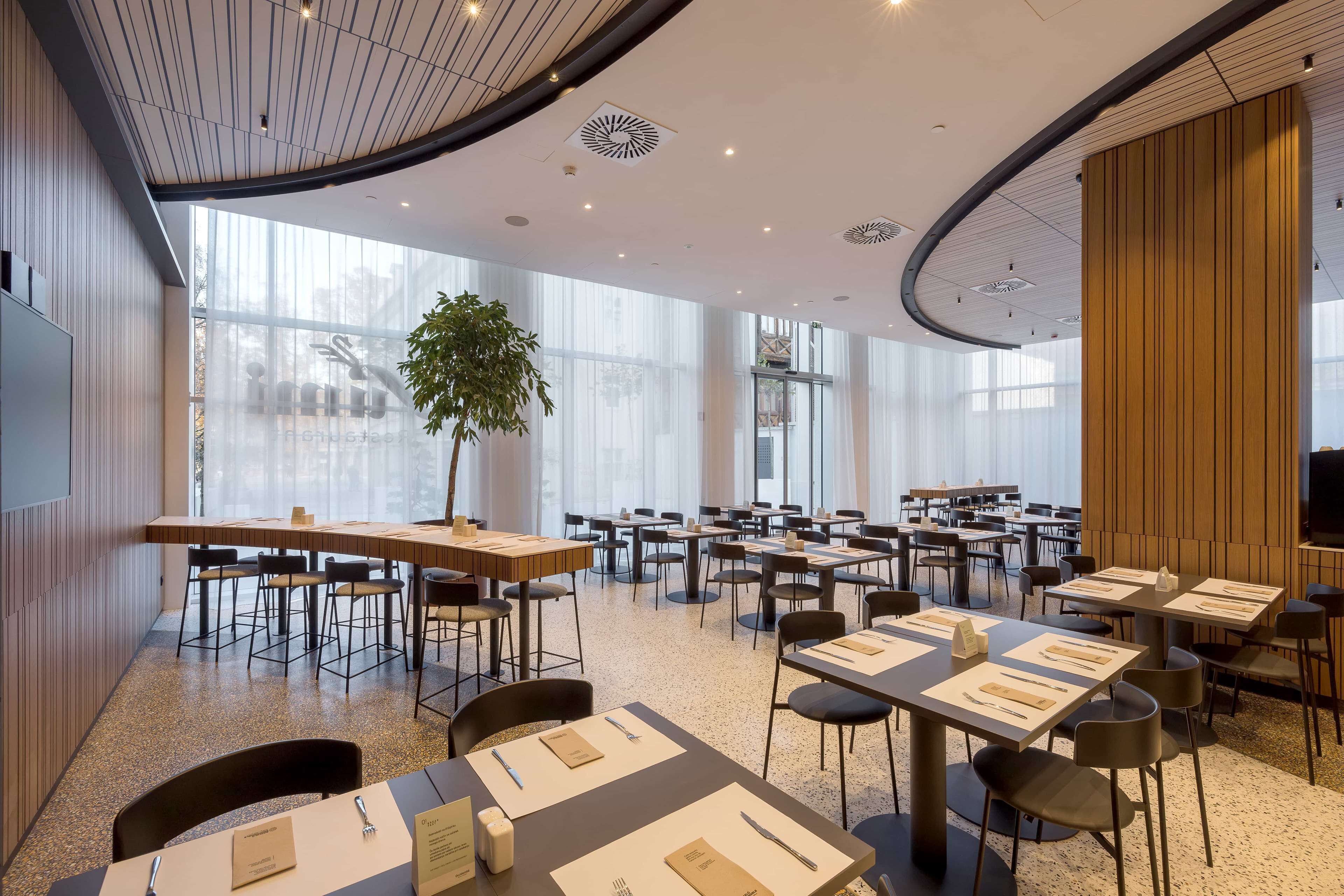 A modern dining area with multiple tables and chairs, large windows, and a central plant.