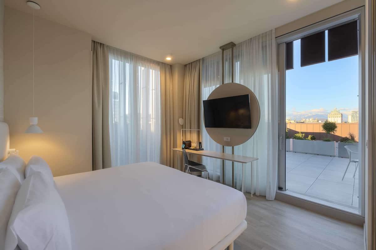 Ein Hotelzimmer mit Bett, Schreibtisch mit Fernseher und Blick auf die Stadt durch große Fenster.