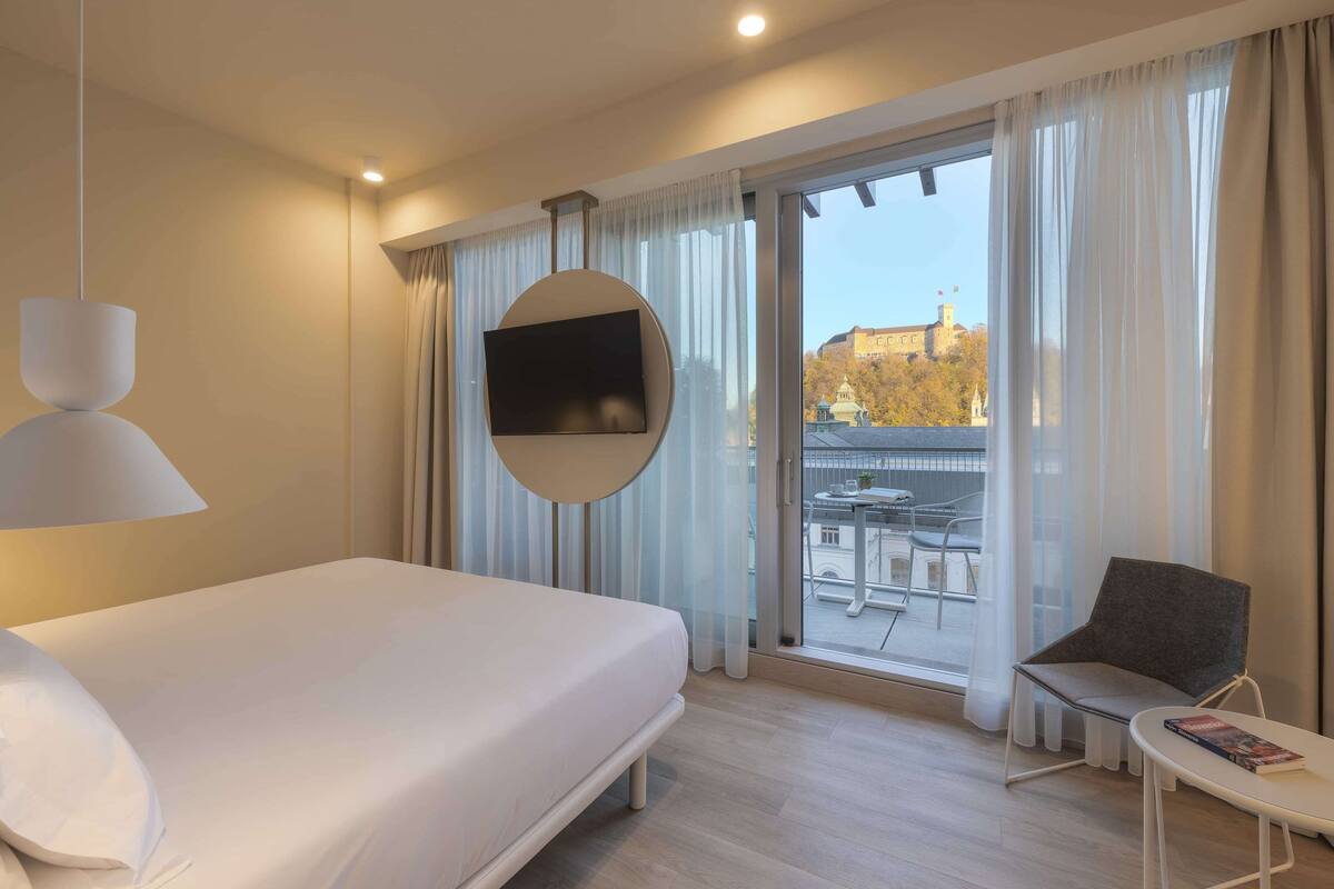 Ein Hotelzimmer mit Bett, Wandfernseher, Balkon mit Tisch und Stühlen sowie Blick auf ein Gebäude mit Bäumen.