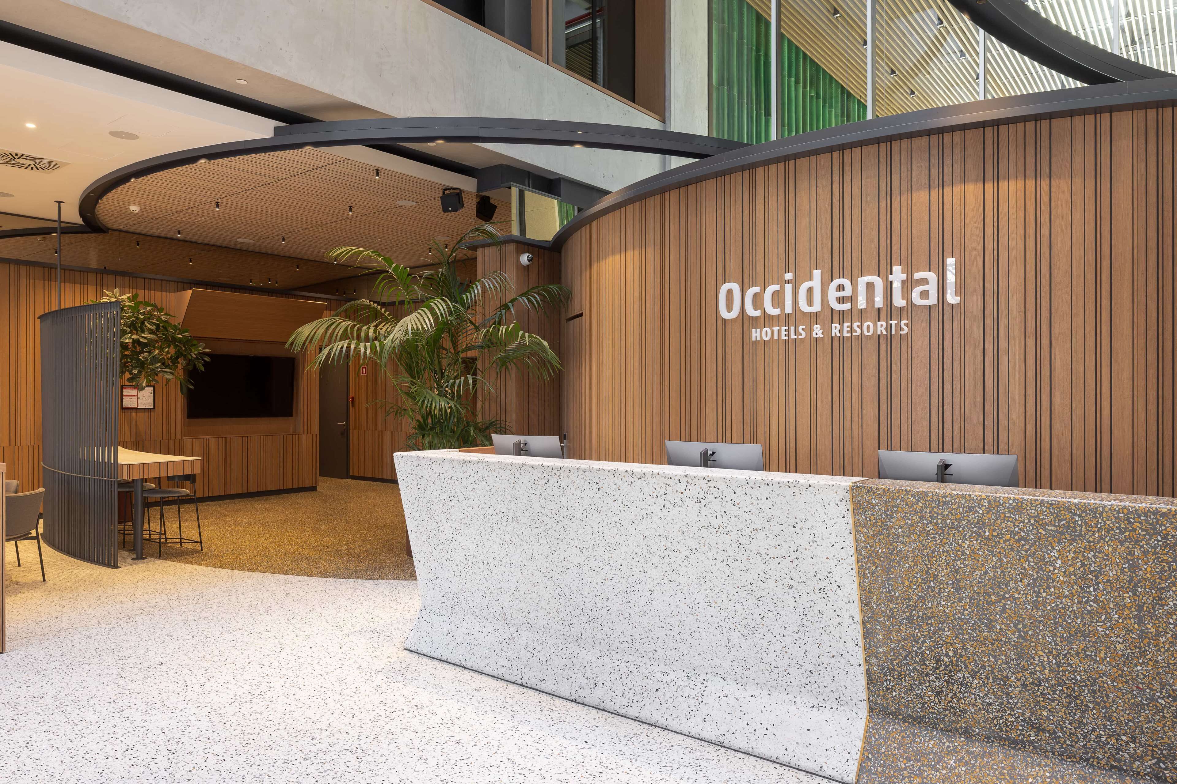 A reception area with a wooden wall and a sign that reads "Occidental Hotels & Resorts." There is a reception desk with a computer and a plant in the background.