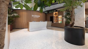 Un espace d’accueil avec un comptoir de réception en bois, un grand arbre en pot et un panneau indiquant « Occidental Hotels & Resorts ».