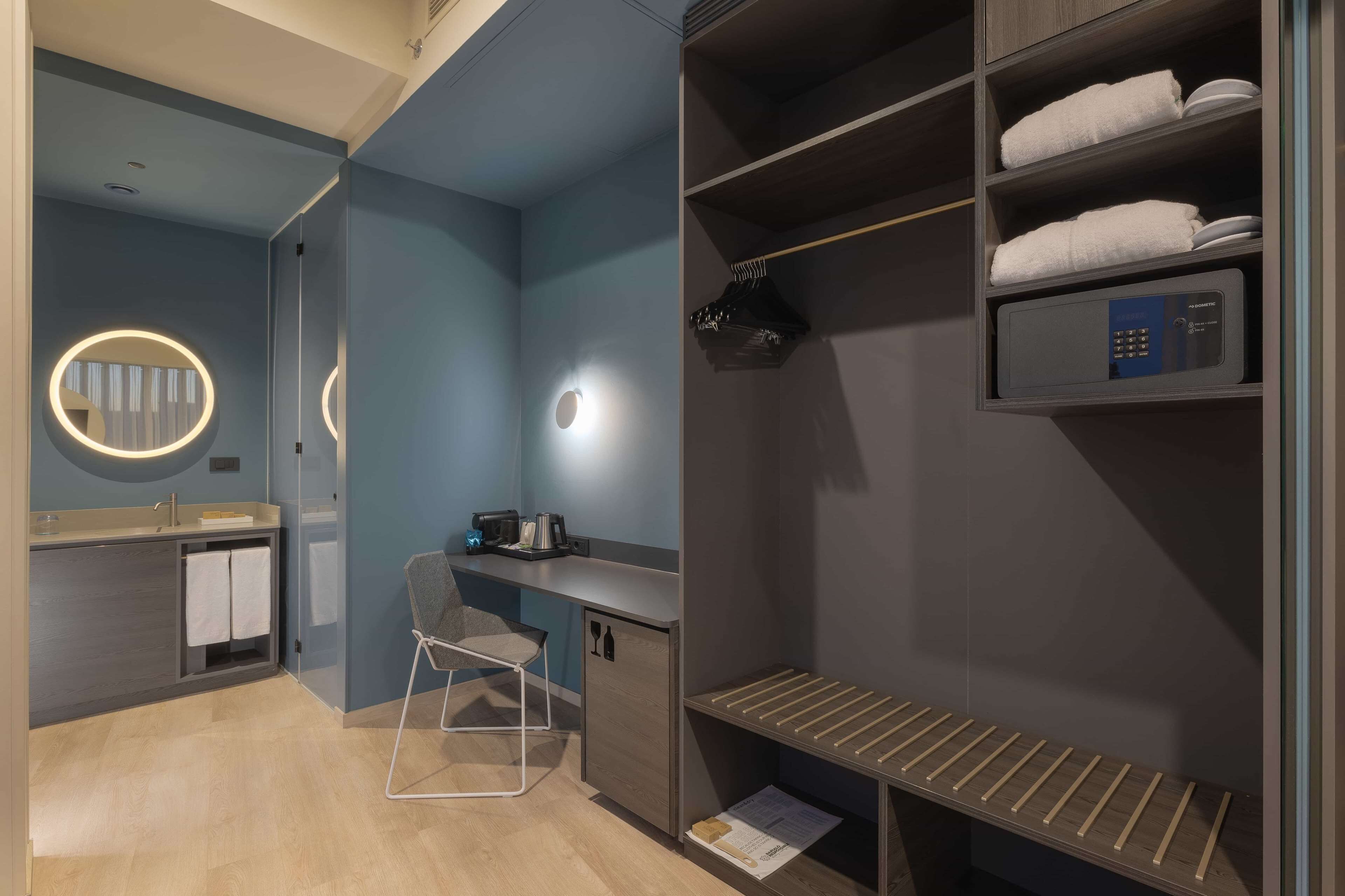 A modern bathroom with a vanity, a mirror with a circular light, a chair, and a built-in heater.