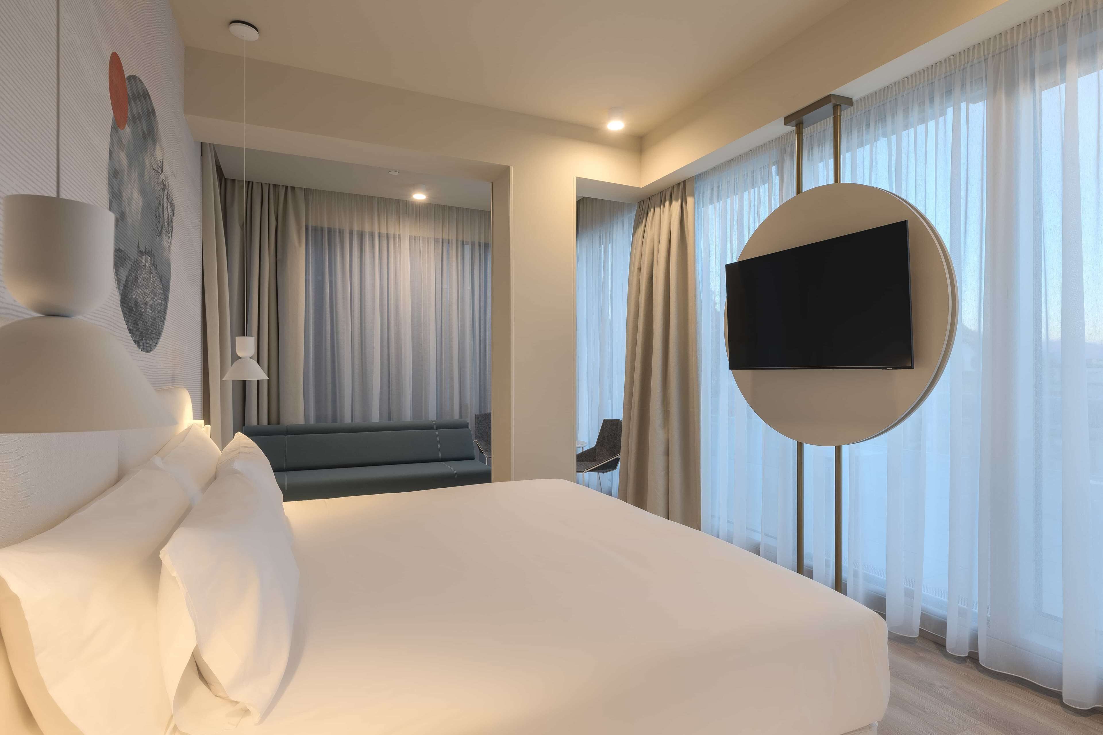 A modern hotel room with a large bed, a flat-screen TV mounted on the wall, and a view of the city through sheer curtains.